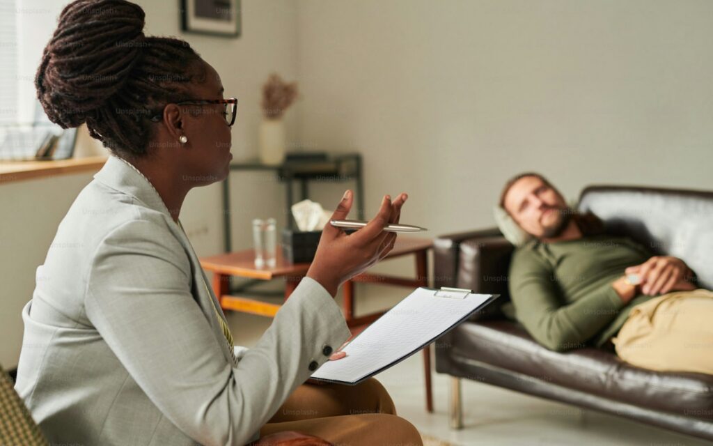 African American psychologist with document sitting on armchair and talking to patient at office